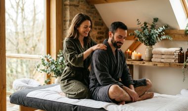 A couple enjoys a shoulder massage on a warmly lit table surrounded by towels and greenery.
